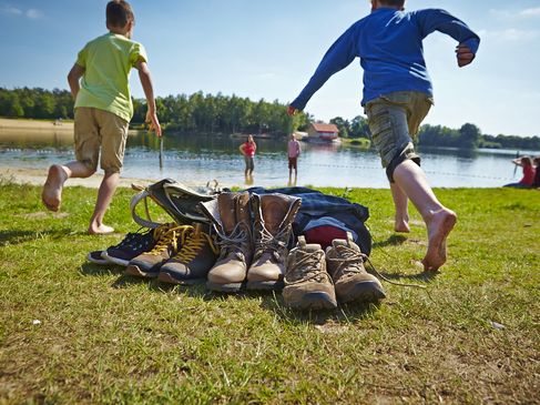 Im Vordergrund stehen die ausgezogenen Wanderstiefel, während links und rechts vom Bildrand Kinder auf den Quendorfer See zulaufen, wo ihre Eltern schon mit hochgekrempelter Wanderhose im Wasser stehen.