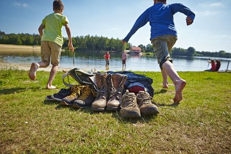 Im Vordergrund stehen die ausgezogenen Wanderstiefel, während links und rechts vom Bildrand Kinder auf den Quendorfer See zulaufen, wo ihre Eltern schon mit hochgekrempelter Wanderhose im Wasser stehen.